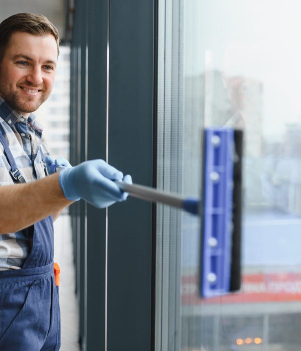 Smiling man in blue gloves washing windows with a squeegee inside an office building, maintaining cleanliness in a modern urban environment